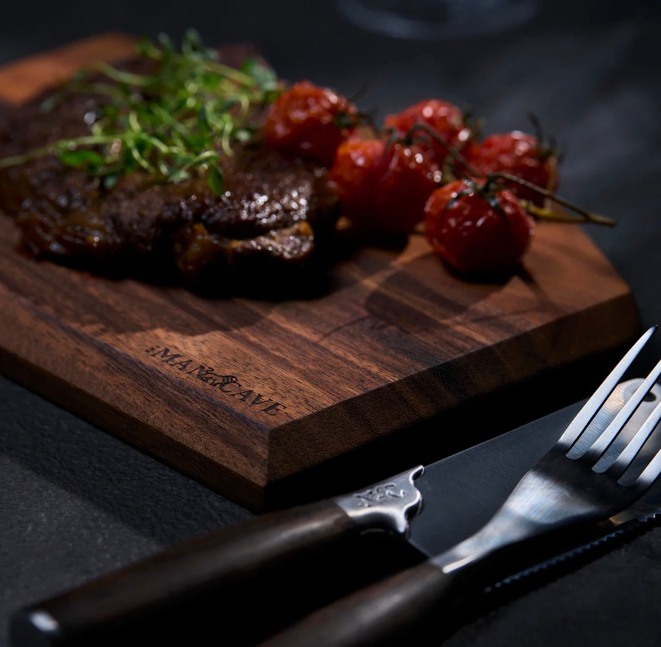 Steak with roasted tomatoes on a wooden cutting board with a knife and fork, branded 'The Mancave'.