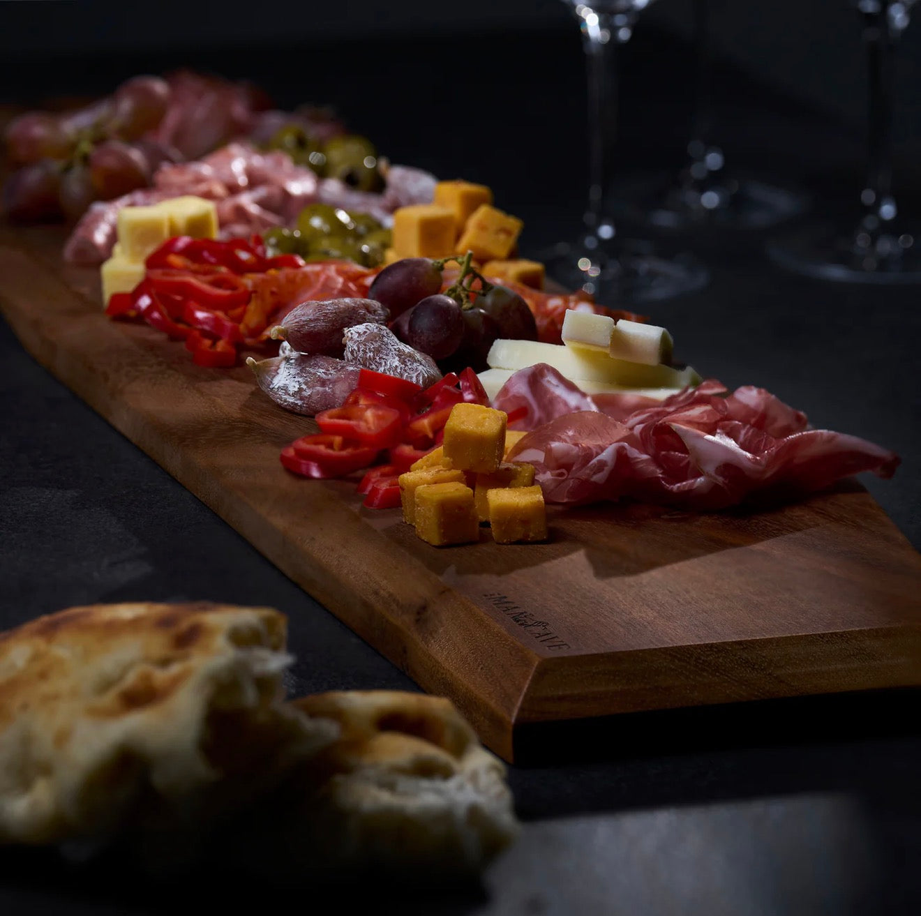 Wooden cutting board with assorted meats, cheeses, and fruits on a dark surface. The Gathering Board
