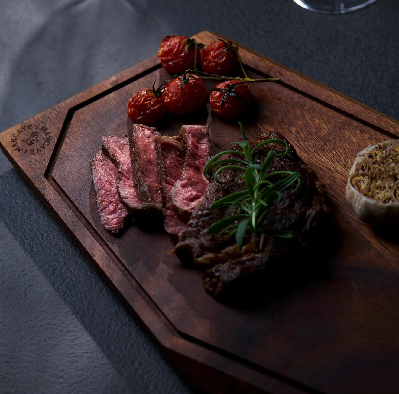 Wooden cutting board with sliced meat, roasted tomatoes, and herbs on a dark surface. The Master´s Cutting Board